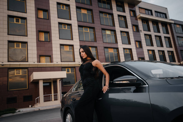 Stylish young girl stands near the car in a black dress. Business fashion and style - Zdjęcie, obraz
