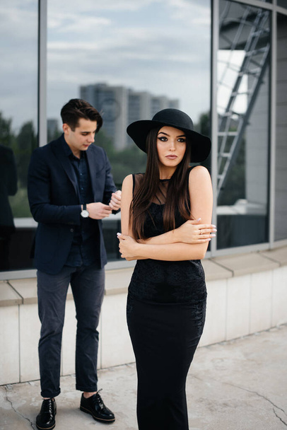 A beautiful, stylish pair of young people in black clothes and glasses stand against the background of an office building in the sunset. Fashion and style - Photo, image