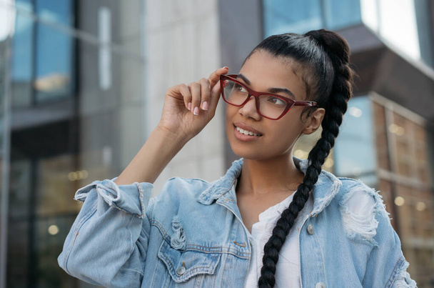 Πορτρέτο της όμορφης Αφρο-Αμερικανίδας που φοράει κομψά γυαλιά, στέκεται στο δρόμο και χαμογελά. Νεαρό χαρούμενο μοντέλο μόδας ποζάρει για φωτογραφίες σε εξωτερικούς χώρους. Έννοια φυσικής ομορφιάς  - Φωτογραφία, εικόνα