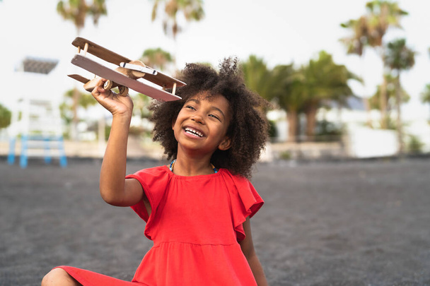 Afro child playing with wood toy airplane on the beach - Little kid having fun during summer holidays - Childhood and travel vacation concept - Photo, Image