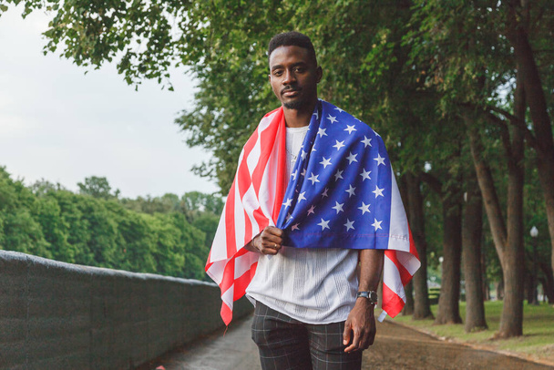 カメラを真剣に見て、屋外に立って、彼の肩に米国の旗を持つハンサムなアフリカ系アメリカ人の男。夏日. - 写真・画像