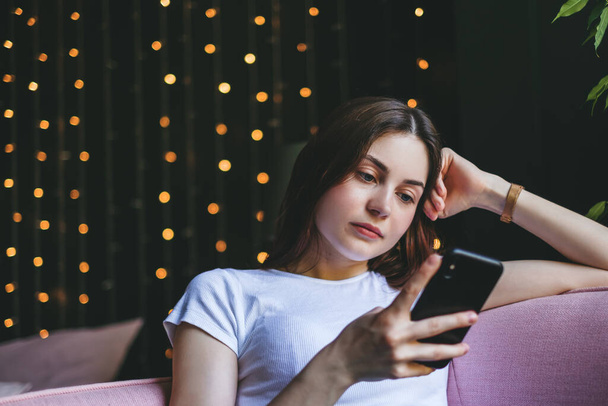 Young pensive caucasian woman use a smartphone while sitting on pink sofa at home. High quality photo - Foto, Imagem