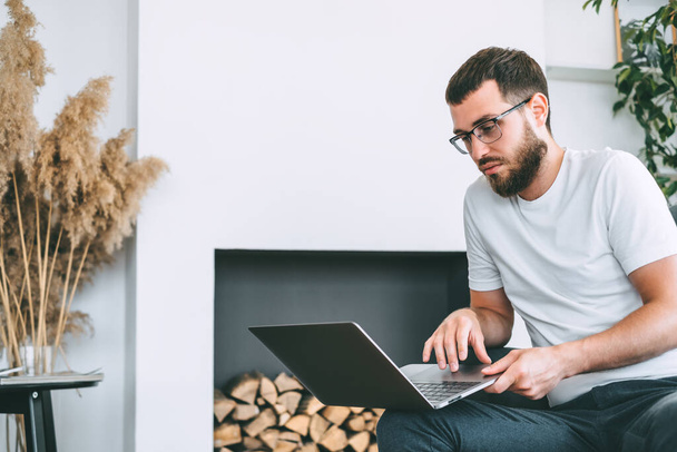 Young caucasian bearded man, freelance worker, sitting on a chair and using laptop computer. Work at home. High quality photo - Foto, immagini