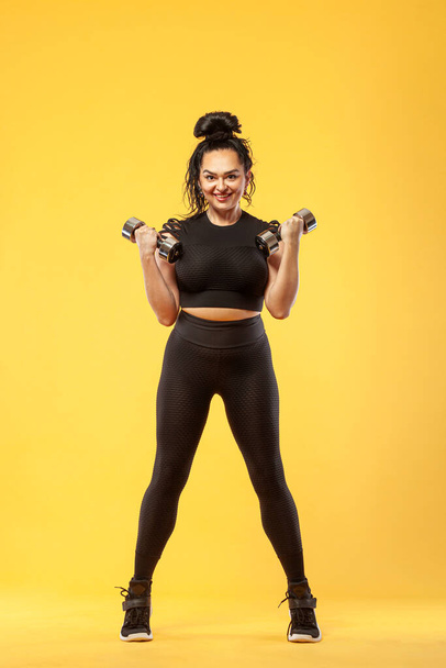 Size plus woman sporty fit woman in black sportswear, athlete with dumbbells makes fitness exercising on yellow background. Motivation for fat people. - 写真・画像
