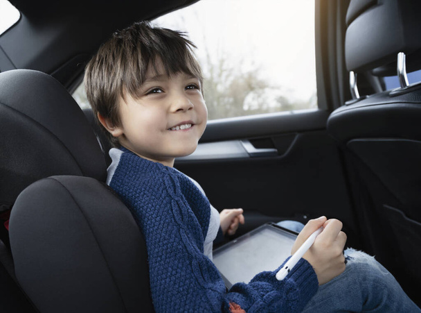 Ευτυχισμένο νεαρό αγόρι χρησιμοποιώντας έναν υπολογιστή tablet, ενώ κάθεται στο πίσω κάθισμα του συνοδηγού ενός αυτοκινήτου με ζώνη ασφαλείας, Παιδί αγόρι σχέδιο για έξυπνο μαξιλάρι, Πορτρέτο του παιδιού διασκεδάζοντας τον εαυτό του σε ένα οδικό ταξίδι.  - Φωτογραφία, εικόνα