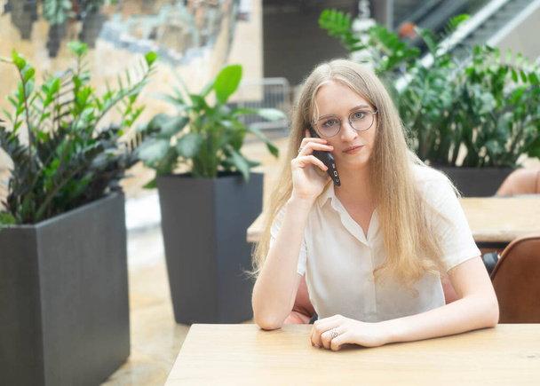 A beautiful cheerful young caucasian blond business woman sitting on the table,talking at phone and looking at camera. Concept of successful young women in a corporation. Girl in a white shirt. Horizontal photo. Blurred background with plants. - Φωτογραφία, εικόνα