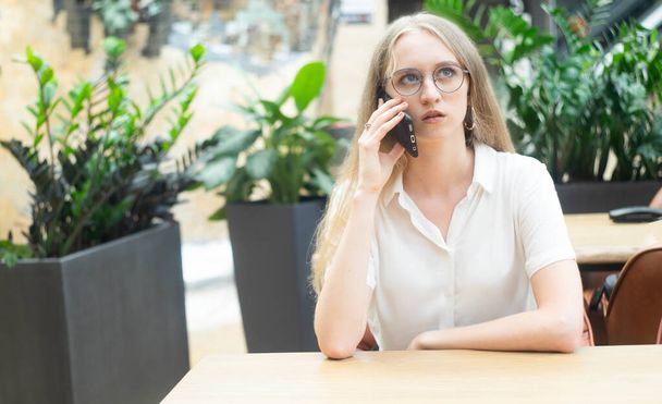 A beautiful cheerful young caucasian blond business woman sitting on the table,talking at phone and looking away. Concept of successful young women in a corporation. Girl in a white shirt. Horizontal photo. Blurred background with plants. - Photo, Image