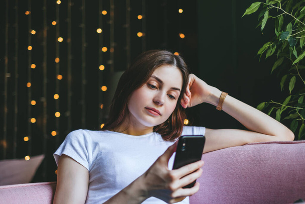 Young pensive caucasian woman use a smartphone while sitting on pink sofa at home. High quality photo - Fotó, kép