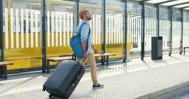 Πίσω από Καυκάσιος νεαρός κομψός άνδρας με καπέλο και σακίδιο με τα πόδια στη στάση λεωφορείου και βαλίτσα με ρόδες. Επιστροφή άποψη για αρσενικό τουριστικό περίπατο σε εξωτερικούς χώρους από το σιδηροδρομικό σταθμό ή το αεροδρόμιο. - Πλάνα, βίντεο