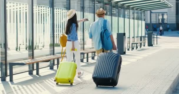 Achter op blanke jonge stijlvolle gelukkige paar reizigers lopen op het busstation en het dragen van koffer op wielen. Achteraanzicht op knappe man en mooie vrouw, toeristen wandelen vanaf treinstation - Video