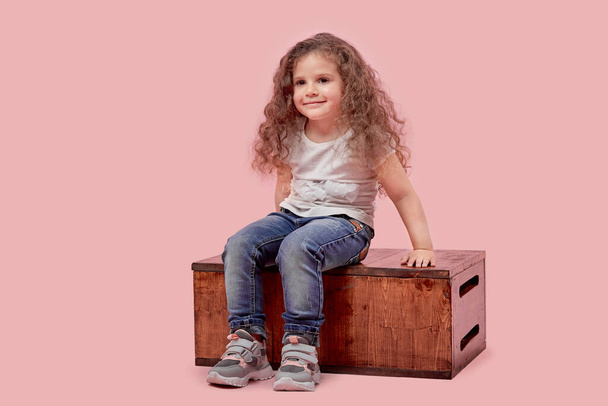 A small child in casual wear sits on a wooden stand on an empty background. - Фото, изображение