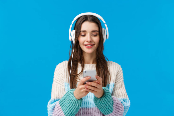 Waist-up portrait smiling, happy good-looking woman with long dark hair, listen music in big headphones, holding smartphone, chatting with friend, send message, pick song, standing blue background - Fotografie, Obrázek