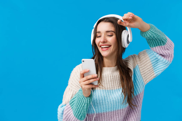 Happy charismatic and carefree smiling european woman enjoying awesome music quality sound in new headphones received as christmas gift, raising hands up holding smartphone, blue background - Photo, image
