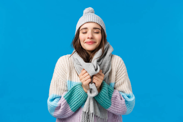 Happiness, dreams and christmas mood concept. Cheerful attractive sensual young woman in winter hat, sweater, touching scarf around neck, close eyes and inhale fresh air, enjoy snowy weather - Photo, Image