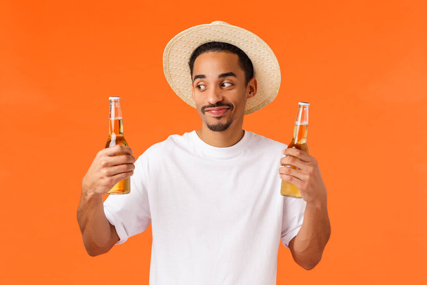 Mutlu, gülümseyen ve şapkalı beyaz adam tropikal bir ülkede dinleniyor, soğuk bir içecekle serinliyor, elinde iki şişe bira tutuyor, turuncu arka planda duruyor. - Fotoğraf, Görsel