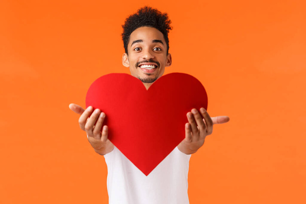 Guy giving his love you. Handsome lovely african-american man stretching hands and presenting red heart, smiling, showing true love and sympahy, express affection, standing orange background - Photo, image