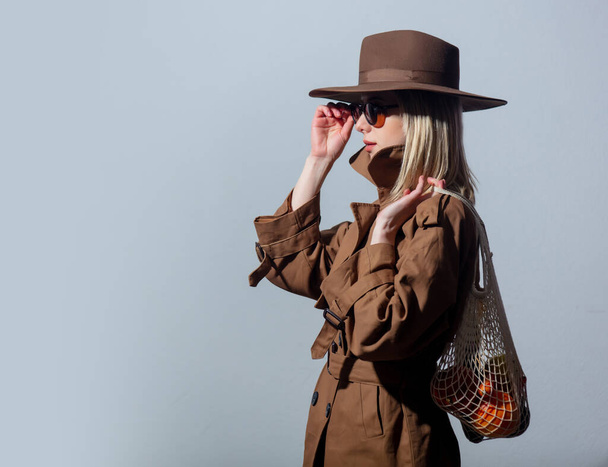 Стильная женщина в солнечных очках и плаще держит чистую сумку на белом фоне
  - Фото, изображение