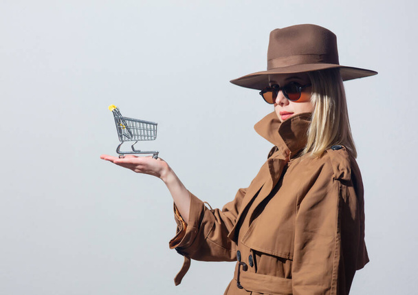 Stilvolle Frau mit Sonnenbrille und Mantel hält Einkaufswagen auf weißem Hintergrund - Foto, Bild