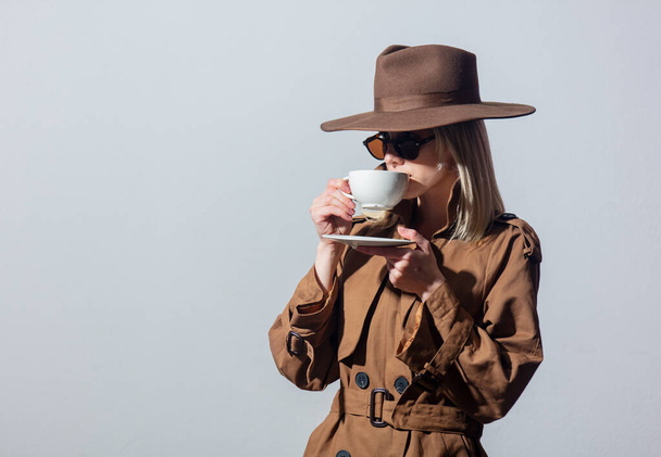 Style woman in cloak, hat and sunglasses holds cup of coffee on grey background - Photo, Image