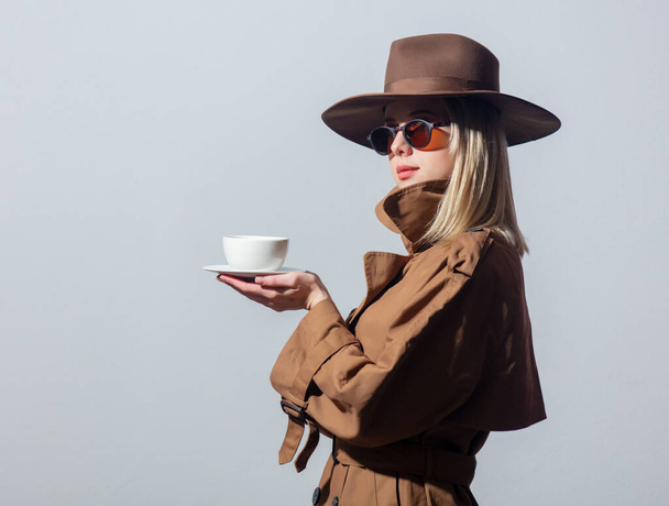 Mujer de estilo en capa, sombrero y gafas de sol sostiene la taza de café sobre fondo gris
 - Foto, imagen