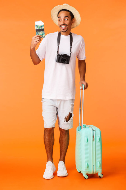 Full-length vertical tiro animado feliz afro-americano viajante masculino, não pode esperar saborear coquetel sob o sol e desfrutar de férias impressionantes, segurando bagagem, mala e passaporte com bilhetes
 - Foto, Imagem