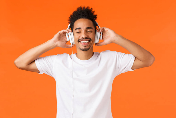 Τεχνολογία, gadgets έννοια. Ευτυχισμένος σύγχρονος αφροαμερικανός άντρας σε λευκό t-shirt με ακουστικά, κλειστά μάτια και χαμογελαστά εκστατικά, ακούστε αγαπημένη μουσική, καλή ποιότητα ήχου, πορτοκαλί φόντο - Φωτογραφία, εικόνα