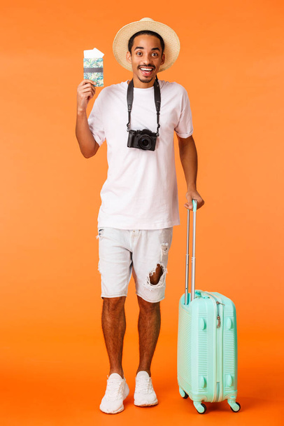 Plein plan vertical afro-américain heureux petit ami a acheté deux billets vacances station de luxe, tenant bagages voyage prêt, terminal de l'aéroport debout avec passeport, fond orange - Photo, image