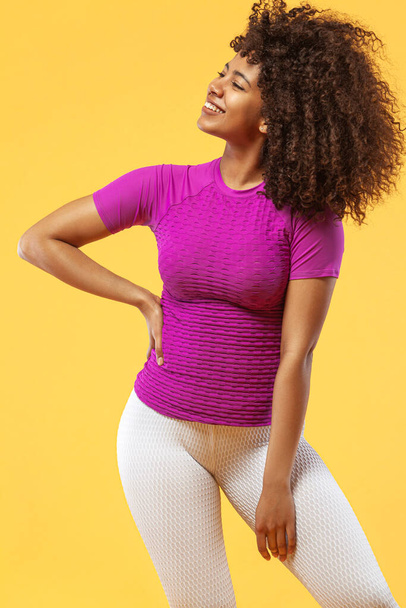 Smiling strong athletic woman with black skin and curly hair, doing exercise on yellow background wearing sportswear. Fitness and sport motivation. - Φωτογραφία, εικόνα