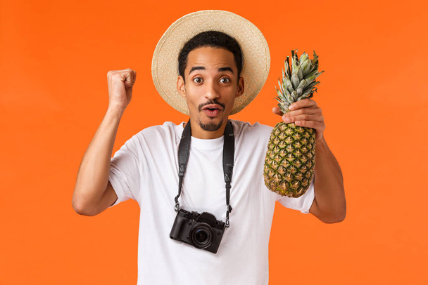 Hooray vacation and tropical fruits. Cheerful excited african-american man finally travel abroad, having fun during trip, fist pump and holding pineapple, standing with camera over orange background - Fotografie, Obrázek