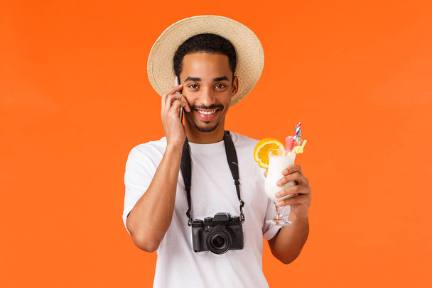 Vakantie, reis en reisbureau concept. Knappe vrolijke charismatische Afrikaans-Amerikaanse man met hoed, camera en wit t-shirt, cocktail drinken, telefoon praten, bellen vanuit het buitenland, oranje achtergrond - Foto, afbeelding