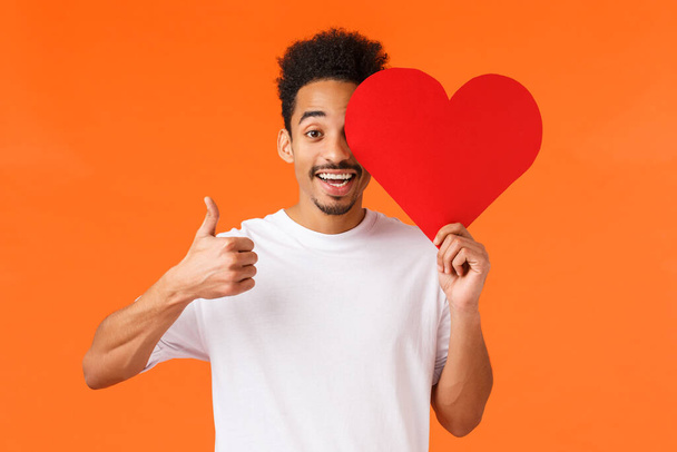 いい子だ。カリスマアフリカ系アメリカ人の笑顔男で白いtシャツ,ロマンチックなバレンタインカードを保持し、親指アップを示します,中に真の愛を見つけました日付アプリ,ボーイフレンドと祝います,オレンジの背景 - 写真・画像