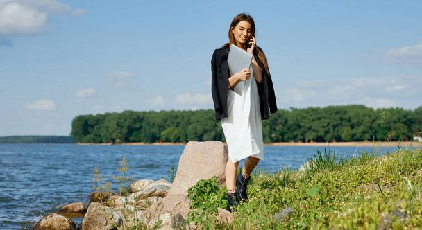 A woman businesswoman walks along the rocky bank talking on the phone and holding a laptop in her hand - Фото, зображення
