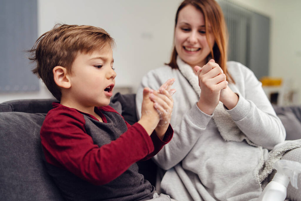 Portrait of small caucasian boy male child kid sitting on the sofa bed at home by mother young woman rubbing hands with alcohol disinfectant antibacterial prevention hygiene product prevent disease - Photo, Image