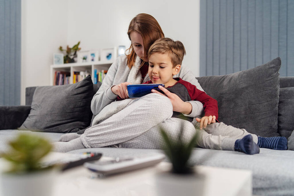 Mother and son young woman and her boy child caucasian kid sitting on the sofa bed at home making video call or playing video games on mobile smart phone during quarantine lockdown - 写真・画像