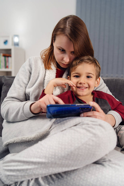Mãe e filho jovem mulher e seu filho menino criança caucasiana sentado no sofá-cama em casa fazendo videochamada ou jogando videogames no telefone inteligente móvel durante a quarentena lockdown vista frontal
 - Foto, Imagem