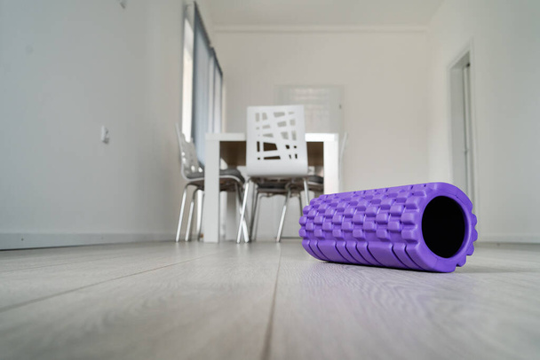 Rodillo de espuma texturizada púrpura para masaje corporal en el suelo de madera blanco claro en casa frente a los muebles borrosos
 - Foto, Imagen