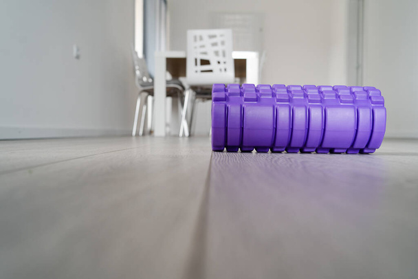 Purple textured foam roller for body massage on the light white wooden floor at home in front of the blurred furniture - Photo, Image