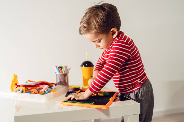 Retrato de vista lateral de un pequeño niño caucásico jugando junto a la mesa en casa sosteniendo el tablero de juguete creativo usando rojo
 - Foto, Imagen