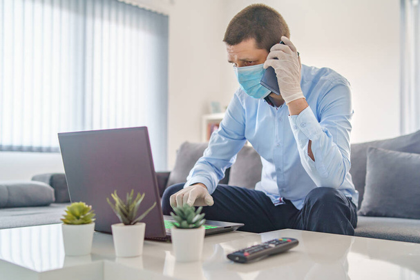 Volwassen blanke man met blauw hemd thuis werkend op laptop, terwijl hij naar een mobiel telefoontje ging met een beschermend gezichtsmasker en handschoenen tijdens een quarantainevirusepidemie. - Foto, afbeelding