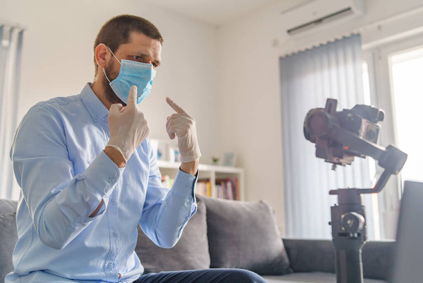 Πλευρική άποψη για Καυκάσιος ενήλικας blogger κάθεται στο σπίτι την ημέρα με κάμερα gimbal σταθεροποιητή κάνοντας βίντεο σχετικά με την εξάπλωση της νόσου του ιού πανδημίας φορώντας προστατευτικά γάντια μάσκας για την πρόληψη της καραντίνας - Φωτογραφία, εικόνα