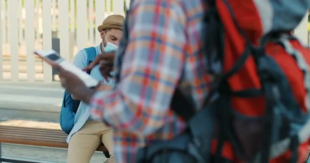 Junges kaukasisches nettes Paar von Reisenden, die auf einer Bank an der Bushaltestelle sitzen und sich unterhalten, während sie nach der Route auf der Landkarte suchen. Sommerferien. Schöner Mann und schöne Frau in medokalen Masken mit Stadtplan. - Filmmaterial, Video