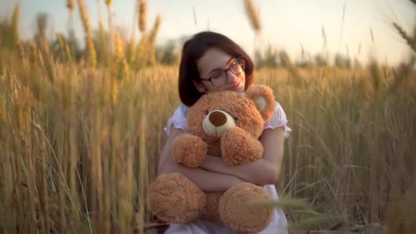 Una giovane donna siede in un campo di grano secco con un orsacchiotto. Ragazza abbraccia un orsacchiotto in mano vista frontale. - Filmati, video