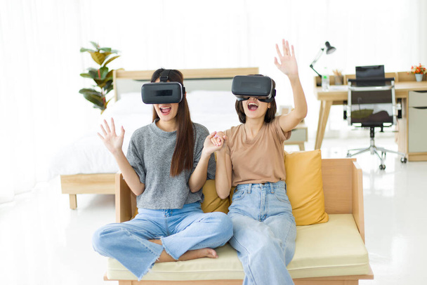 Chicas adolescentes que son amigas íntimas que usan gafas VR mientras están sentadas en el sofá, dos chicas asiáticas jóvenes están sorprendidas y emocionadas con la realidad virtual que vio.
. - Foto, Imagen