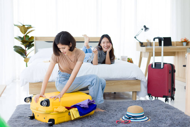 Giovane donna asiatica preparazione viaggio valigia a casa mentre la sua amica sdraiata sul letto e ridevano di lui a causa di troppi vestiti. - Foto, immagini