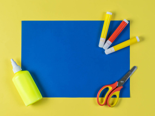 Blaues Blatt Papier, Klebstoff, Filzstifte und Scheren auf gelbem Hintergrund. - Foto, Bild