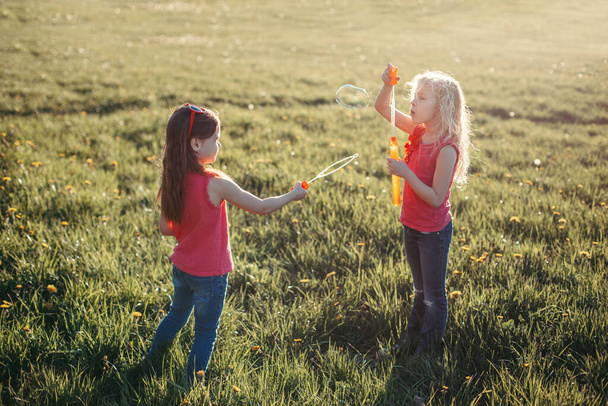 Attrape une bulle. Filles amis soufflant des bulles de savon dans le parc le jour d'été. Les enfants s'amusent dehors. Authentique moment magique d'enfance heureuse. Mode de vie activité saisonnière pour les enfants. - Photo, image