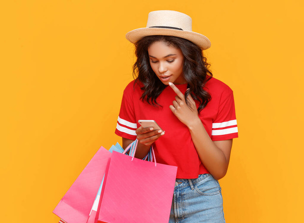 Κομψό έθνικ θηλυκό με χάρτινες σακούλες σκάλισμα πηγούνι και περιήγηση προσφορές έκπτωσης στο smartphone κατά τη διάρκεια ψώνια κατά κίτρινο backgroun - Φωτογραφία, εικόνα
