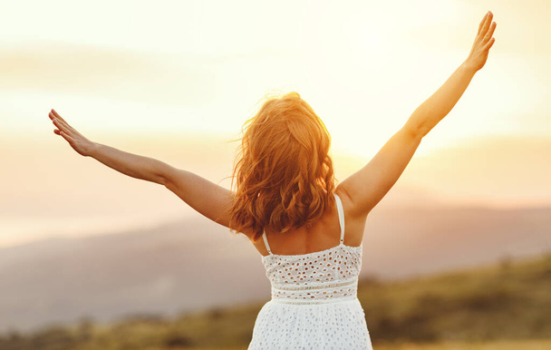 Вид ззаду на невідому молоду жінку в білому платті, що піднімає руки і насолоджується свободою, стоячи на зеленому лузі на розмитих пагорбах і заході сонця під час літніх канікул
 - Фото, зображення
