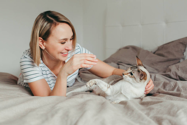 Porträt einer glücklichen kaukasischen lächelnden blonden Frau, die zu Hause im Schlafzimmer auf dem Bett liegt und mit einer orientalischen, punktfarbenen Katze spielt. Haustierbesitzer mit Haustier.  - Foto, Bild
