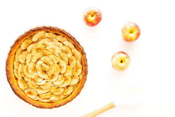 Zelfgebakken Franse appeltaart, een open appeltaart, in een witte keramische schaal naast de Gala appels en een witte kooklepel op een witte achtergrond. Vlakke lay, bovenaanzicht. - Foto, afbeelding